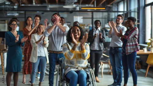 A group of business people are clapping and celebrating with one woman in a wheelchair. The modern office space shows a diverse group of employees standing around lady in the wheelchair cheering her