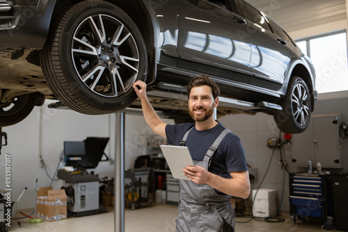 Smiling mechanic using tablet in auto repair shop with car on lift. Confident male technician diagnosing vehicle problem using modern technology. Professional car service in well-equipped garage.