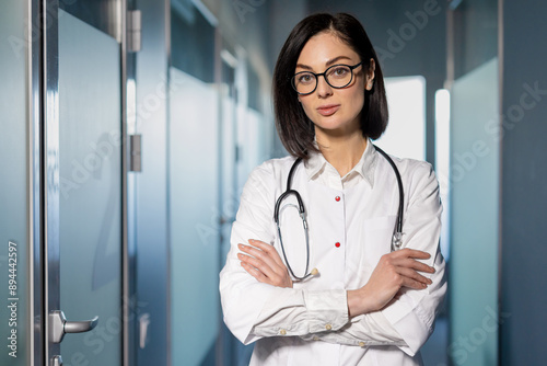 Confident female doctor with glasses and stethoscope standing in hospital corridor. Professional medical staff in healthcare environment. Concept of medical expertise, healthcare, and hospital work.