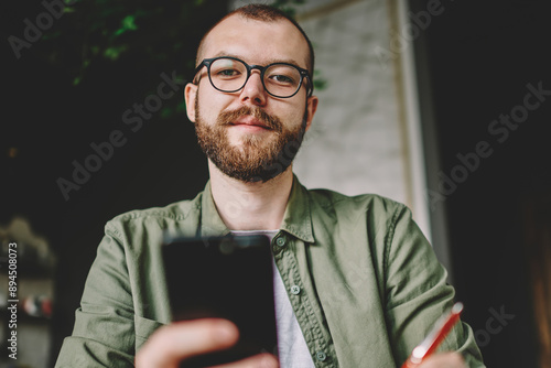 Below portrait of bearded hipster student in stylish eyeglasses for vision correction holding modern smartphone in hands and rewriting information from internet website while looking at camera