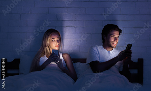 Cheating Concept. Unfaithful Husband Chatting On Cellphone Lying With Wife In Bed At Night. Selective Focus, Low Light