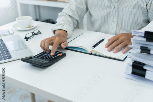 office employee works on desk, using calculator to manage financial documents and accounts, preparing an income tax report in a busy office setting.