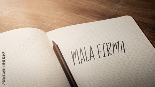 A handwritten inscription "Mała Firma" on a grille of an open notebook on a wooden countertop, next to a black pencil, lighting of light. (selective focus), translation: Small Company