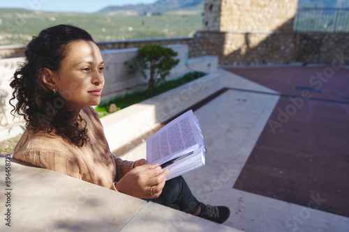 Young woman sitting outdoors reading a book, enjoying serene surroundings on a sunny day