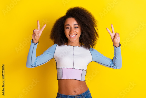 Photo of cheerful optimistic girl with perming coiffure dressed stylish shirt showing v-sign symbol isolated on yellow color background