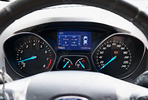 Close-up of a car dashboard with illuminated gauges and digital display showing various vehicle metrics. The image focuses on the speedometer, tachometer, fuel gauge.