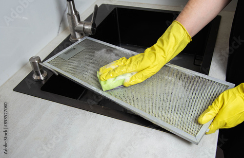 A girl's hands in yellow gloves with a sponge wash the kitchen hood filter with detergent to remove grease and dirt. Cleaning the kitchen, grease stains