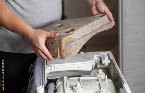 The master holds the counterweight stone of the washing machine in his hands. The concept of replacing the counterweight due to vibration and noise when spinning a washing machine, industry