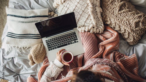 Woman comfortably shopping online with a laptop in bed, surrounded by pillows and a cup of tea 