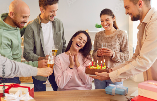 Happy friends group congratulating a young smiling girl sitting at table on her birthday giving her cake and present gift boxes at evening party at home. Happy birthday and celebration concept.