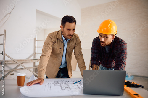 Construction worker and home owner using laptop while analyzing blueprints.