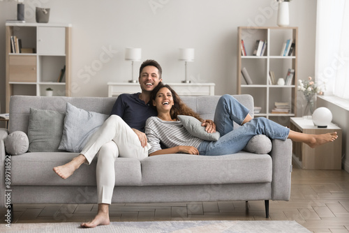 Cheerful couple spend leisure time at home, relaxing on cozy sofa, looking at camera, pose, smiling, feel happy about moving to new apartment, enjoy warm relations. Tenants family, renters portrait