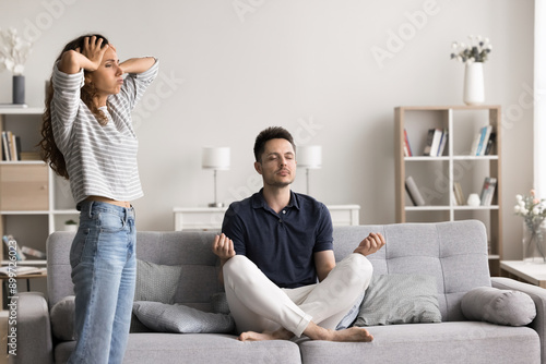 Frustrated or stressed woman, holding her head with her hands, while her husband sit on couch in lotus position, practicing meditation with eyes closed. Relationships problems, hysterics, drama queen