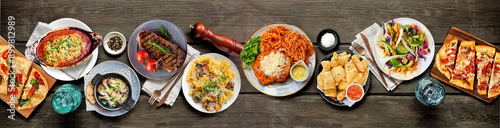 Table scene of various delicious foods. Above view on a dark wood banner background. Pastas, flatbreads, tacos, steak and restaurant theme dishes.