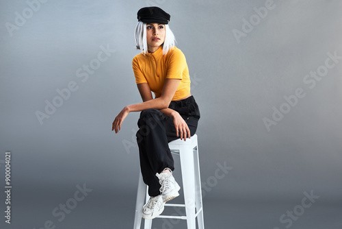 Woman, stool and fashion in studio with hat for confidence, thinking and style inspiration ideas. Model girl, chair and designer clothes with newsboy cap, thoughts and streetwear by gray background