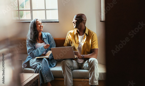 Happy colleagues laughing and collaborating while sitting on a couch with a laptop in a modern office