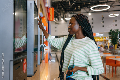 Black Woman with glasses and striped shirt using a touch screen kiosk in a modern shopping mall. Girl chooses burger on touch menu of fast food restaurant. 