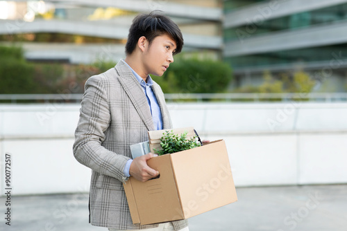 A young Asian man walks away from a modern office building, carrying a cardboard box filled with personal items. He appears dejected and downtrodden, perhaps having just been laid off or resigned