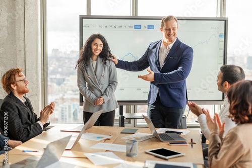 A group of business professionals gathered around a conference table in a modern office space are applauding a young lady colleague who has just finished giving a presentation