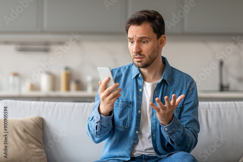 A man sits on a light gray couch in a kitchen, staring at his smartphone with a confused expression. He has his right hand raised, gesturing with his fingers, while holding the phone in his left hand.