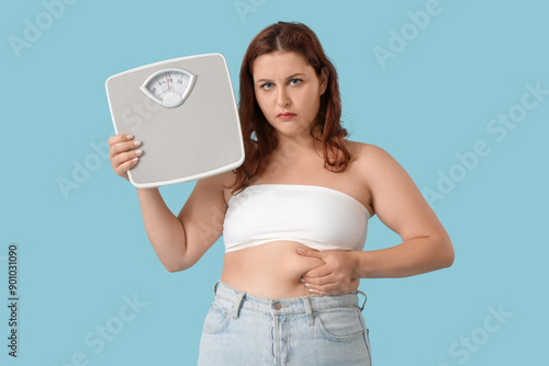 Overweight young sad woman with scales on blue background. Weight gain concept