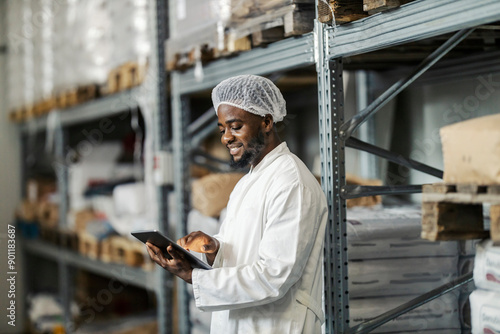 Side view of happy diverse food factory supervisor standing in warehouse and tracking goods and shipment on tablet.