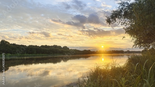 On an early summer morning, the sun rose over the forest on the far side of the river, breaking through the clouds and the fog billowing over the water. Grass, bushes and trees grow on the banks