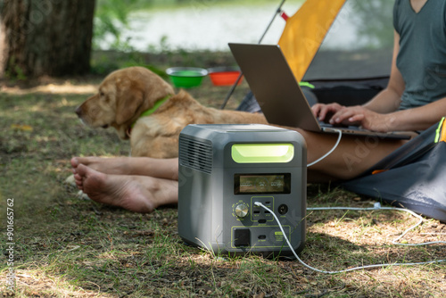 Female digital nomad uses portable power station to charge her laptop during workation on the wild nature. Her dog laying beside.