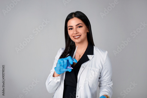 Portrait of an American beautiful female cosmetologist doctor in a white coat with an injection syringe. Medical student. Female hospital worker looking at camera and smiling, studio, gray background