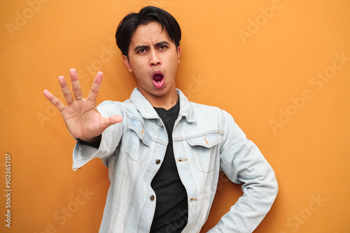 Asian young man with serious face posing making stop sign with hand, Angry Asian young man doing stop gesture isolated on yellow background, defensive gesture, suitable for advertising purposes.