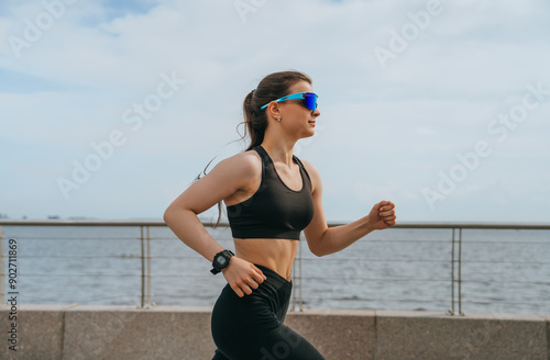 young woman in black sportswear jogging by the water, wearing sunglasses and a watch. The focus is on her upper body, showing her in mid-stride against a blue sky