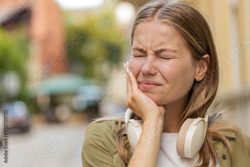 Portrait of young Caucasian woman standing outdoors feeling toothache bad unpleasant symptoms suffering from pain holding hand on cheek jaw damaged dental nerve needs medical help. Girl on city street