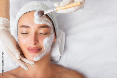 Facial beauty treatment. Beautiful woman getting mask at spa salon, cosmetologist applying white mask with brush, top view, free space