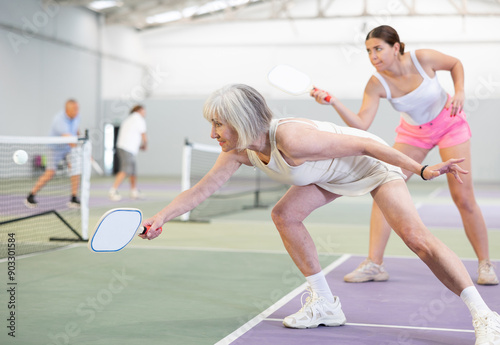 Portrait of positive fit elderly woman playing pickleball on indoor court, swinging paddle to return ball over net ..