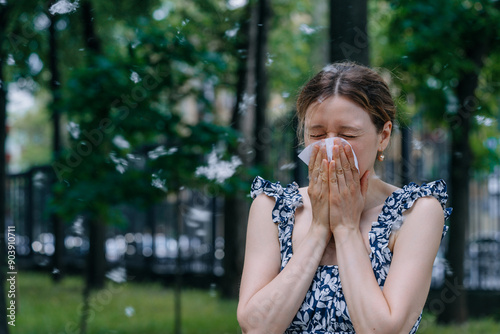 Woman sneezes into a tissue while standing outside in a park, surrounded by airborne allergens like poplar fluff.