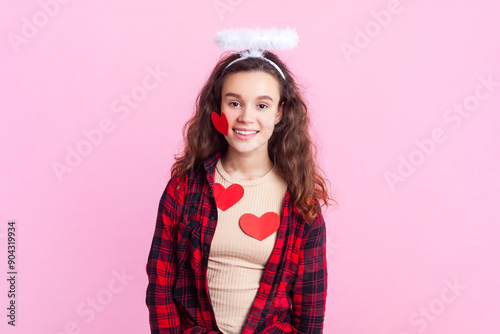 Portrait of romantic flirting teenage girl with wavy hair in red checkered shirt and nimb over head looking at camera with smile covered with red hearts. Indoor studio shot isolated on pink background