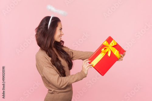 Side view portrait of angelic charming woman with wavy hair and nimb over head giving present box, wearing wearing brown pullover. Indoor studio shot isolated on pink background