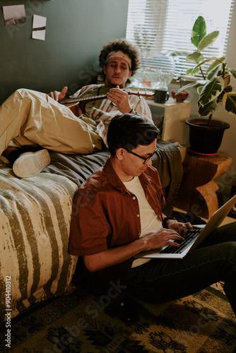 Best friends resting after lectures in dorm room, curly-haired student playing guitar while his friend playing computer games