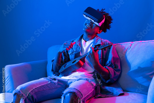 Young African American singer using through VR glasses enjoy singing song with guitar playing on meta blue neon light wall virtual reality metaverse fantasy technology sitting on sofa. Contrivance.