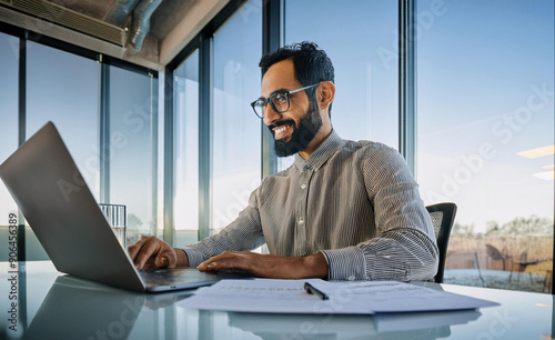 A smiling young man in a suit sits in the office at a desk and works on a laptop, typing on a keyboard, texting, working with data