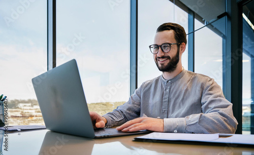 A smiling young man in a suit sits in the office at a desk and works on a laptop, typing on a keyboard, texting, working with data