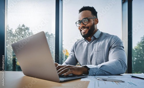 A smiling young man in a suit sits in the office at a desk and works on a laptop, typing on a keyboard, texting, working with data