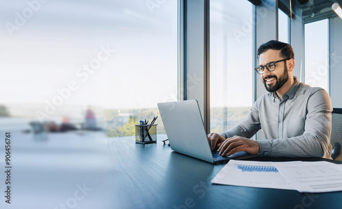 A smiling young man in a suit sits in the office at a desk and works on a laptop, typing on a keyboard, texting, working with data