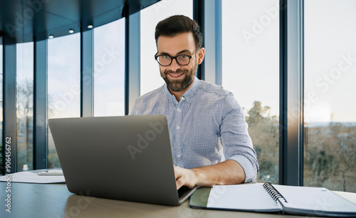 A smiling young man in a suit sits in the office at a desk and works on a laptop, typing on a keyboard, texting, working with data