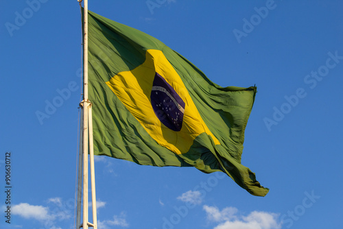 Close up em bandeira do Brasil hasteada balançando ao vento com céu azul ao fundo.