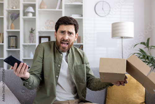 Frustrated man holding smartphone and open delivery box expressing disappointment. Emotional reaction to unsatisfactory online shopping experience, customer dissatisfaction delivery problem at home.