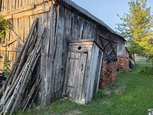 An old, weathered wooden door with rusty metal hinges and a diamond-shaped cutout, attached to a rustic shed or outhouse in a rural setting. The surrounding wood is aged and worn, with grass and trees