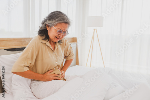 Elderly Asian woman had stomach pain with pained expression on her face while sleeping on the bed, holding her stomach with her hands in excruciating pain for no apparent reason.