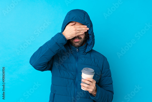 Man wearing winter jacket and holding a takeaway coffee over isolated blue background covering eyes by hands. Do not want to see something