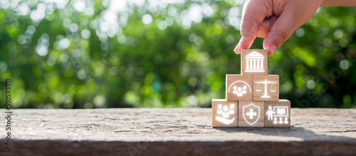 Hand stacking wooden blocks with icons symbolizing social policy elements like healthcare, justice, and community support on a natural background.